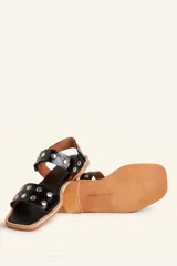 Sandalia baja de cuero vacuno color negro con acabado efecto charol texturizado, decorada con tachas metálicas plateadas en las tiras y la sujección del talón. Cuenta con suela plana de cuero.