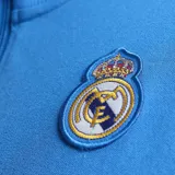 Campera deportiva Adidas del Real Madrid, modelo Terrace Icons. Color celeste con detalles en blanco y dorado. Presenta cierre frontal, cuello alto, bolsillos laterales, logo de Adidas y escudo del Real Madrid bordados.