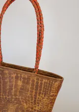 Cartera tipo tote de gamuza color marrón, con correas trenzadas y detalle de piedras naturales incrustadas en líneas verticales. Cuenta con cierre metálico y forro interior de tela.