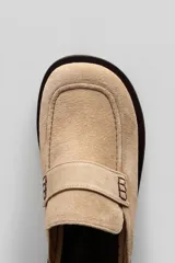 Mule tipo mocasín de gamuza en color tostado, con detalle de costuras decorativas y suela plana de goma negra.