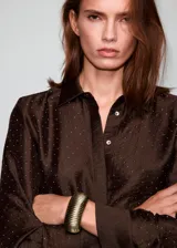Camisa de satén color marrón chocolate, con cuello camisero, cierre frontal de botones ocultos y mangas largas con puños abotonados. Presenta un diseño recto adornado con pequeños detalles de strass brillantes distribuidos por toda la prenda.