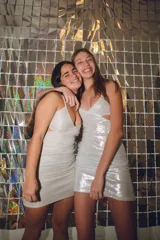 Vestido corto de fiesta color plateado con acabado brillante. Presenta un diseño de dos piezas que simula un conjunto de top y pollera, con aberturas laterales en la cintura, escote en V y tirantes finos que se cruzan en la espalda.