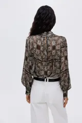 Blusa de tejido transparente con cuello elevado y botones forrados en la nuca. Presenta mangas largas con puños abotonados y un bajo levemente redondeado con estampado paisley.