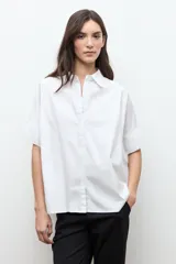 Camisa blanca de corte oversized, confeccionada en algodón, con cuello clásico, cierre frontal de botones y mangas cortas con vuelta.