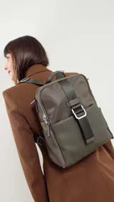 Mochila de tamaño mediano, color beige claro, con cuerpo principal de tela impermeable y detalles en simil cuero liso en la parte inferior y correas. Presenta un asa superior corta y correas ajustables para llevar al hombro o cruzada. Cuenta con un bolsillo frontal con cierre y un detalle decorativo de correa con hebilla metálica rectangular.