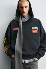 Sudadera tipo hoodie de corte relajado, color negro, con capucha y bolsillo frontal tipo canguro. Presenta parches decorativos en el pecho y estampados en color amarillo en la manga derecha. Acabados en rib en puños y ruedo.
