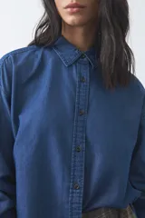 Camisa de jean en tencel con silueta amplia, hombros caídos, cuello clásico y cierre frontal con botones. Presenta ruedo curvo con tajos laterales y pliegue en la espalda.