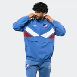 Buzo tipo rompevientos de nylon con medio cierre frontal, cuello alto y puños elásticos. Presenta un diseño en bloques de color azul, blanco y bordó, con el escudo del Club Nacional de Football y el logo de Umbro estampados en el pecho.
