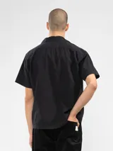 Camisa negra de manga corta con cuello camp y calce relajado. Confeccionada en una mezcla de lino y algodón, terminada con botones de madera y parche característico de Polonio. Hecha en Italia.