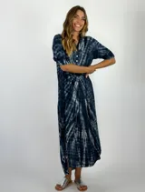Vestido camisero midi con estampado tie-dye en tonos azul y blanco. Presenta cuello camisero, cierre frontal parcial con botones y mangas tres cuartos con trabillas. El diseño es holgado y cuenta con un ruedo asimétrico, más largo en la parte posterior.