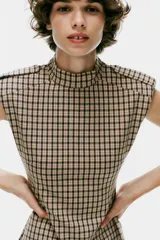 Mono corto de cuello subido y manga sisa con hombreras, con diseño de cuadros tipo tartán en tonos beige y negro. Cuenta con trabillas en los hombros y cierre en la espalda con cremallera oculta.