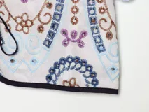 Remera corta de poliéster color crudo con bordados en tonos azules, marrones y morados, escote en V con ribete oscuro y cierre frontal con cordones y cuentas.