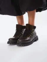 Botas acordonadas de estilo montañés, confeccionadas en cuero marrón oscuro con detalles en gamuza al tono. Presentan una suela track robusta de goma negra con costura visible y ganchos metálicos para los cordones.