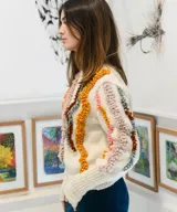 Sweater tejido a mano en punto jersey de 100% lana merino, color blanco con detalles texturizados en punto bucle de tonos rosados, mostaza, marrón y gris, formando líneas onduladas en el frente y las mangas. Cuenta con cuello redondo base y puños largos acANALados.