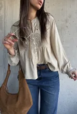 Camisa beige de manga larga con detalles bordados en el frente, cuello redondo y cierre frontal con botones. Las mangas presentan puños con volados.