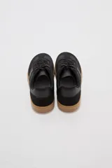 Zapatillas urbanas de color negro con suela de goma marrón, cordones negros con detalles blancos y paneles laterales.