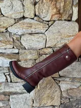 Bota de caña alta estilo biker, confeccionada en cuero color chocolate. Presenta un cierre metálico lateral que recorre toda la caña, estribo y cadena decorativa desmontables, y taco cuadrado con plataforma.