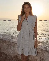 Vestido corto de lino color gris claro, sin mangas, con volados en los hombros, cuello mao y cierre frontal con botones de nácar. Presenta una falda con vuelo y detalle de volado en el ruedo.