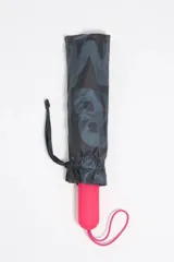 Paraguas plegable con funda textil estampada con el logo de Bimba y Lola en tonos oscuros (negro/gris). El mango es de goma en color fucsia vibrante con el nombre de la marca grabado.
