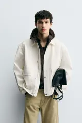 Cazadora de hombre, corte relaxed fit, color blanco hueso o crema, con acabado brillante. Presenta cuello solapa, manga larga, dos bolsillos de plastrón con solapa en la cadera y cierre frontal con botones a presión ocultos por una solapa.