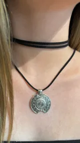 Gargantilla doble de gamuza negra, compuesta por un choker ajustado al cuello y un collar largo con dije circular de metal plateado con diseño de espiral y detalles grabados.