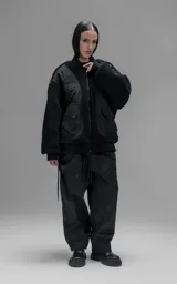 Campera estilo bomber unisex, confeccionada en una combinación de corderito sintético y paneles de ripstop. Presenta un diseño utilitario con múltiples bolsillos frontales con cierres y broches a presión, cuello, puños y ruedo en ribb elástico.