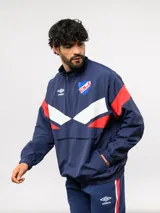 Buzo deportivo de nylon con medio cierre frontal y cuello alto. Presenta un diseño de bloques de color en azul, blanco y bordó, con el escudo del Club Nacional de Football y el logo de Umbro en el pecho.