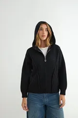 Abrigo negro con capucha, confeccionado en tejido de textura suave. Presenta cierre frontal con cremallera, bolsillos laterales integrados con costuras decorativas y puños ajustados.