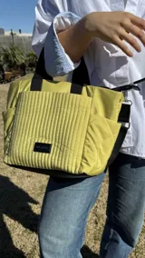 Cartera tipo tote de nylon color malva, con diseño capitoneado de líneas verticales en el frente y rombos en los laterales. Cuenta con doble asa de mano en color negro, bolsillos laterales y un parche decorativo con logo en la parte frontal.