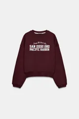 Sudadera de cuello redondo y manga larga, color bordó, con estampado frontal en blanco que dice "Chasing The Sunset's Glow SAN DIEGO 1980 PACIFIC HARBOR".