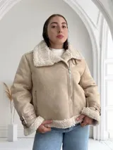 Chaqueta color beige con interior de piel sintética blanca, cierre cruzado con cremallera y detalles de hebillas en los puños.