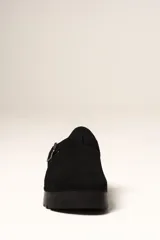 Zueco de plataforma color negro, con capellada de gamuza sintética y detalle de hebilla metálica ajustable en el empeine. Presenta una base de corcho en el talón y suela dentada de goma.