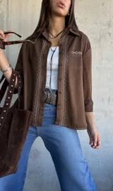 Camisa de gamuza color chocolate con bordados decorativos en color crudo a lo largo de la botonadura frontal y en el bolsillo superior. Presenta cuello clásico, cierre frontal con botones y mangas largas.
