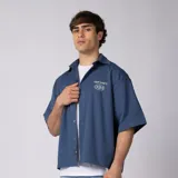 Camisa de manga corta azul oscuro de corte boxy, confeccionada en algodón. Presenta cuello camisero, cierre frontal con botones y un pequeño bordado en el pecho con las siglas 'VCP CNPT' y el número '333'.