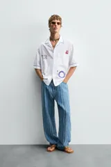 Camisa blanca de algodón con cuello bowling y manga corta, corte relajado. Presenta bordados a contraste en azul, rojo y amarillo que simulan elementos de cocina y artesanía, además de ribetes de pespunte azul en cuello, puños y abertura frontal.