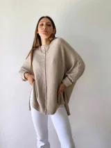 Buzo de tejido de punto color visón, con diseño holgado, cuello redondo, mangas largas con puños acanalados y costura central vertical visible. Presenta un ruedo asimétrico con aberturas laterales.