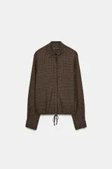 Blusa de la colección Zara Woman, confeccionada en hilatura de viscosa con estampado de cuadro vichy en tonos marrón y negro. Presenta cuello solapa, manga larga con puño y bajo ajustable con cordones. Cierre frontal con botones.
