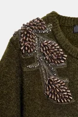 Chaqueta de punto color verde oliva, con cuello redondo, manga larga y cierre frontal con botones. Presenta detalles de abalorios bordados en los hombros.