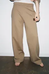 Pantalón de felpa color beige, tiro medio, con cintura elástica, bolsillos laterales ocultos y pierna ancha. Presenta un parche pequeño con un logo en la parte delantera.