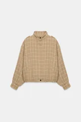 Chaqueta bomber de color beige con diseño a cuadros blancos, cuello subido, manga larga con trabillas en los hombros, bolsillos delanteros y bajo con elástico. Cierre frontal con cremallera y botones a presión.