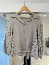 Camisa de lino color beige, con cuello camisero, mangas abullonadas y laterales con detalle de bordado inglés y ojales. Presenta cierre frontal con botones y tiras para anudar en la parte inferior delantera.
