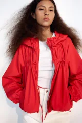 Chaqueta corta de nylon color beige, con cuello subido, capucha y cierre frontal con cremallera. Presenta mangas largas con puños elásticos, bolsillos laterales y detalles de cordones ajustables con lazadas en el pecho y el bajo.