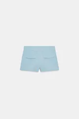 Shorts de tiro medio color crema, con bolsillos laterales y falsos bolsillos con solapa en la parte trasera. Cierre frontal con cremallera y ganchos metálicos.