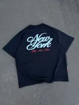 Remera negra de corte oversize con estampado gráfico en la espalda con la inscripción 'New York' en blanco y detalles en rojo, y diseño frontal minimalista.