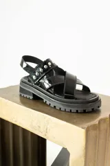 Sandalias de plataforma estilo track, color negro, confeccionadas en material sintético efecto charol. Presentan tiras cruzadas en el empeine y una tira trasera con detalle de tachas metálicas plateadas.