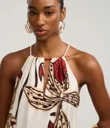 Vestido midi de corte evasé, con escote halter y abertura frontal en el busto, anudado en la parte superior. Presenta un estampado grande de flores y hojas en tonos rojo oscuro, beige y negro sobre fondo crudo. El diseño incluye un volante fruncido en el ruedo inferior.