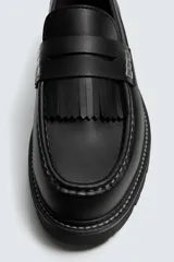 Mocasín negro de cuero liso con antifaz y flecos en el empeine. Presenta costuras blancas visibles y suela track gruesa de goma.