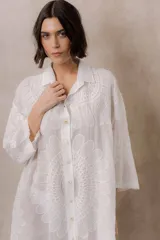 Camisola blanca de silueta amplia y caída liviana, con diseño clásico de cuello camisero, cierre frontal con botones y mangas tres cuartos, decorada con grandes motivos florales bordados.
