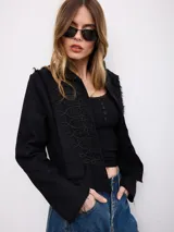 Chaqueta corta negra de gabardina con detalles de cordones decorativos bordados en el frente y bordes deshilachados.