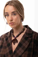 Collar corto con cuentas cilíndricas en tonos marrones y dije central grande en forma de hoja de color marrón oscuro. Presenta detalles de pequeñas cuentas en azul y rojo en la unión del dije con el collar.